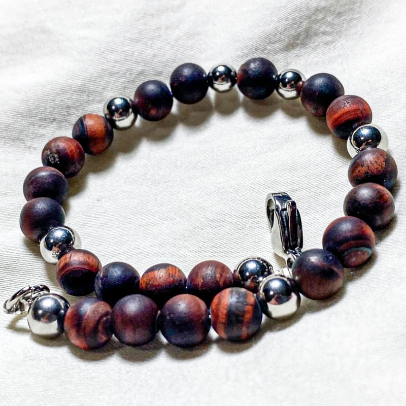 Bracciale in pietra naturale opaca di Occhio Tigre Rosso e acciaio Linea Gimo_Jewels Realizzato con pietre dure di Occhio di Tigre Rosso, con chiusura a moschettone e distanziatori in acciaio anallergico. Pietra originaria del Sud Africa estratta tuttavia anche in Australia, India, Brasile e Birmania, presenta colori e sfumature molto particolari tanto da essere ampiamente utilizzata in gioielleria. Non s9i tratta di un minerale particolarmente pregiato e questo fa sì che sia rarissimo trovarne in giro di contraffatti. In generale si presenta di colore giallo brunastro con striature scure, ma ne esistono anche colori che virano dal blu al grigio al verde (occhio di falco) e dal colore rossastro o mogano (occhio di tigre rosso).